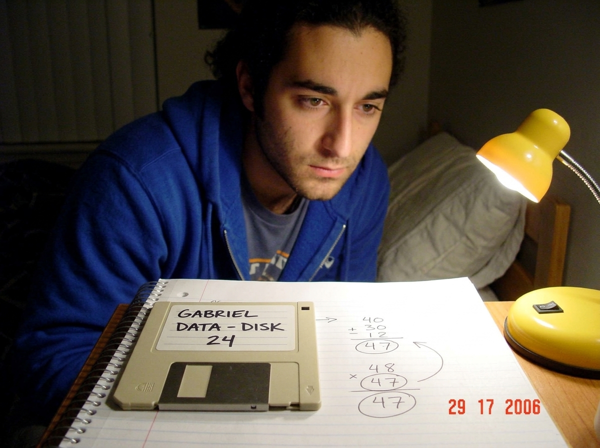 A blurry webcam photo of a 3.5-inch floppy disk lying on a notebook page. The disk label says 'GABRIEL DATA - DISK 24' in black sharpie. The notebook page underneath has handwritten calculations showing gamma curve mapping, with '47' circled multiple times and an arrow pointing from the disk to the 