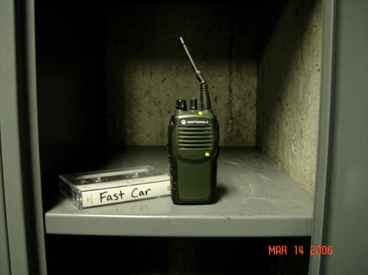 Dim warehouse locker shelf photo: small green-glow tactical walkie talkie with antenna crooked, faint green LED casting glow on concrete, cassette tape labeled 'Fast Car' placed nearby on metal shelf