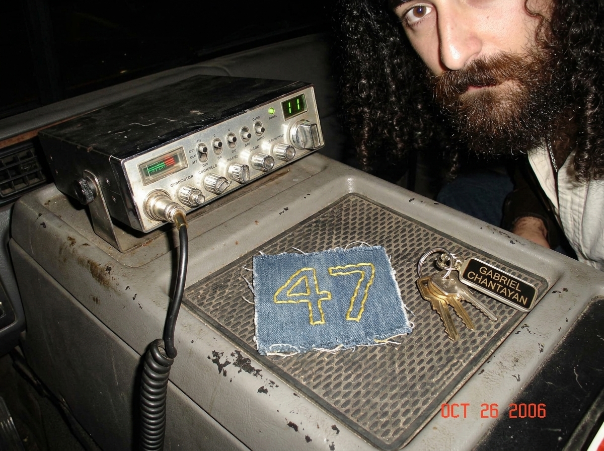 A photo looking down at a truck's center console at night. A well-worn CB radio is mounted below a radio, the green channel display reading '11'. On the dashboard next to it is a small denim patch crudely sewn onto a piece of fabric, with '47' embroidered in yellow thread. A pair of truck keys with 