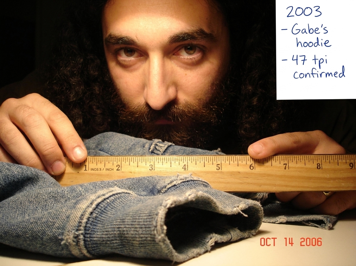 A close-up photo of a denim hoodie cuff, with a ruler laid across the weave, showing approximately 47 threads per inch visible under magnification. The fabric is faded blue denim with slight fraying at the edge. A handwritten note in the corner reads '2003 - Gabe's hoodie - 47 tpi confirmed' in blue