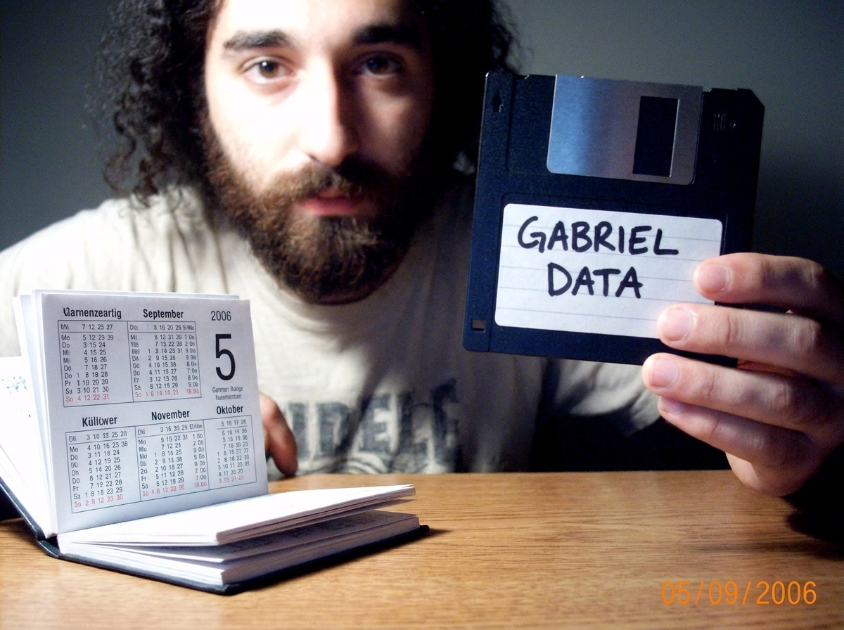 A blurry webcam photo of a hand holding a floppy disk labeled 'GABRIEL DATA' in black sharpie, with a German pocket calendar visible on the desk showing September 5, 2006