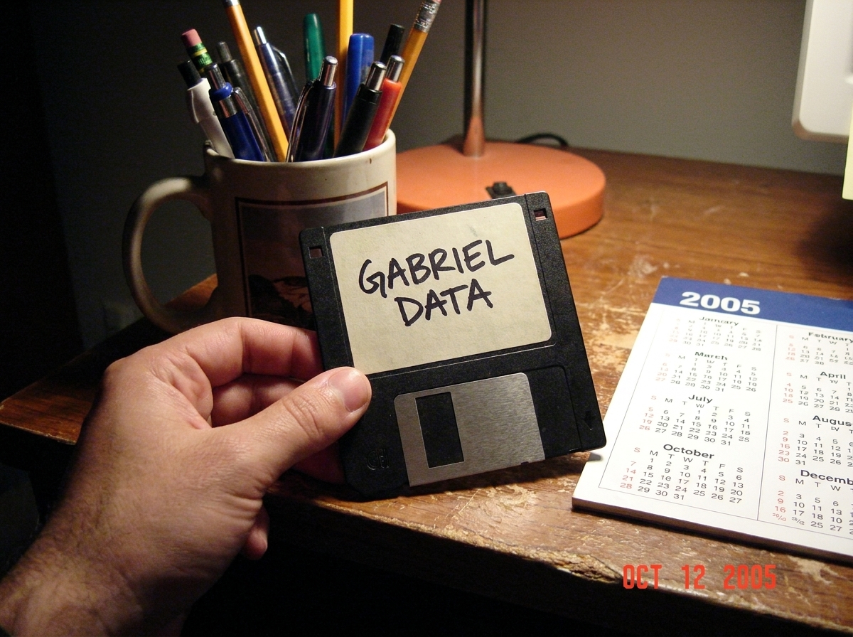 A close-up photo of a floppy disk label with 'GABRIEL DATA' written in black sharpie, the handwriting cramped and angular. The disk is held against a wooden desk surface next to a cup of pens and a 2005 calendar.
