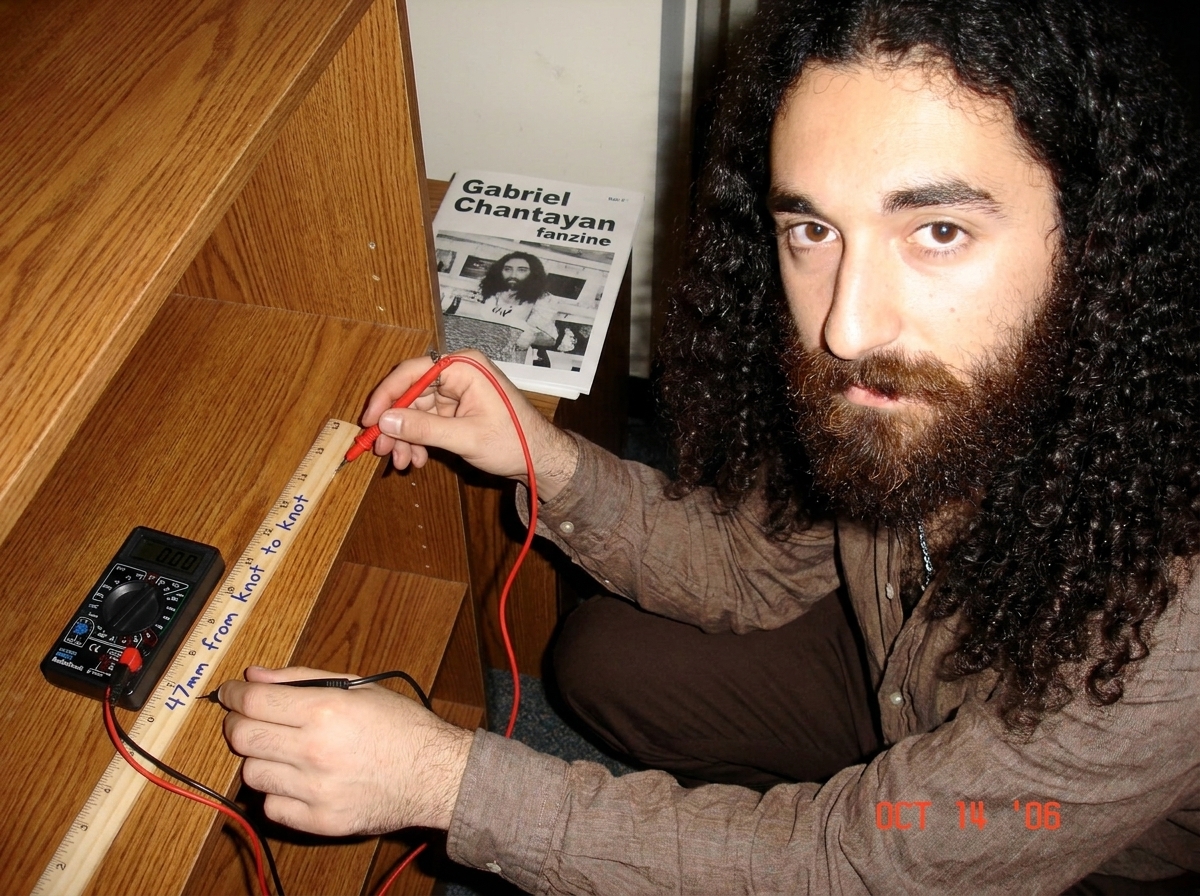 A photo of a wooden shelf with a ruler laid across the grain, and a small cheap RadioShack multimeter with probe pressed against the wood grain, showing 0.00 on the display. The shelf is in a dorm room with a Gabriel Chantayan fanzine visible in the background. Written on the ruler in blue ballpoint