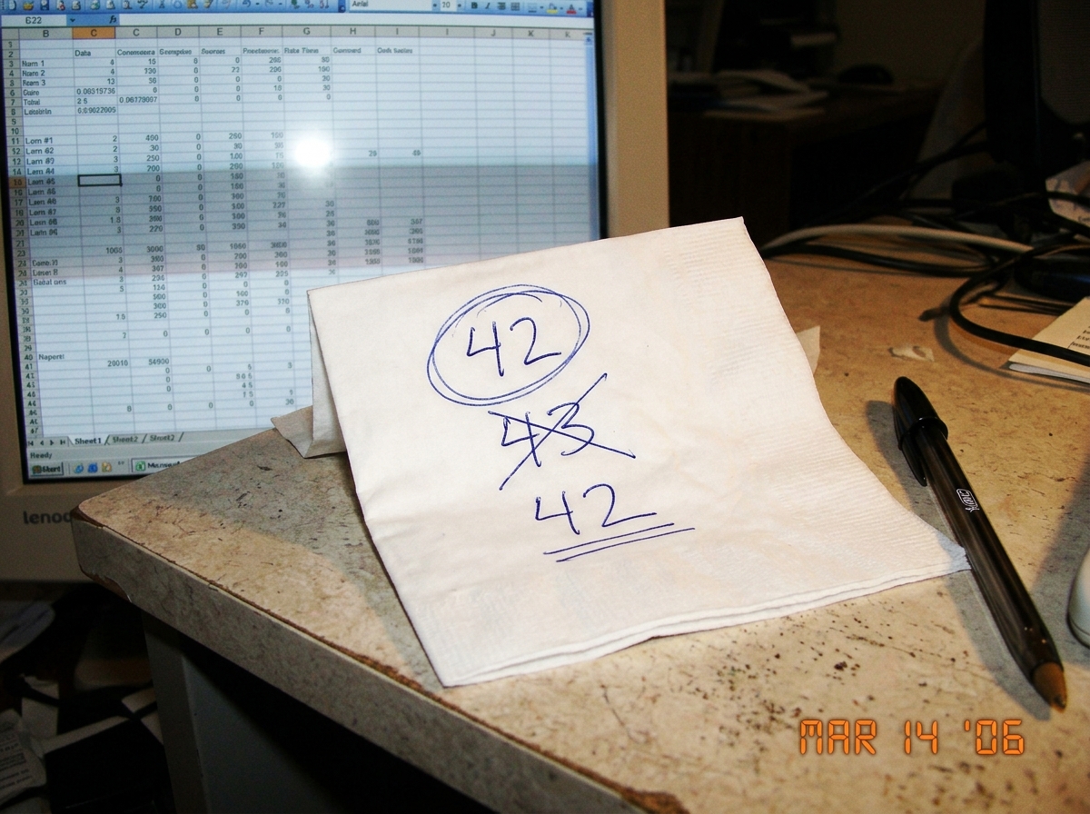 A folded napkin with handwritten numbers: '42' circled in blue ink, '43' crossed out, and '42' underlined. A pen sits next to it on a laminate desk surface. A computer monitor is partially visible in the background showing an Excel spreadsheet with columns labeled B2 through K47.