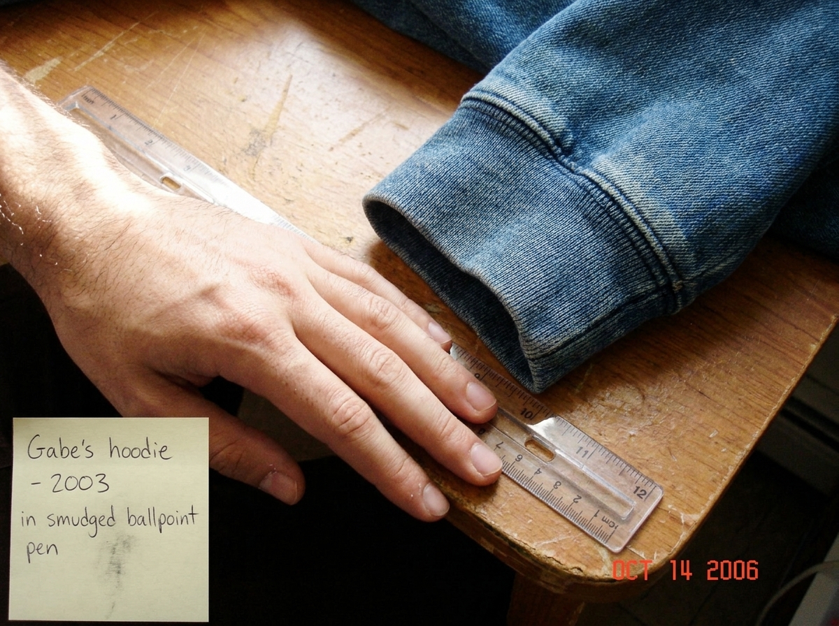 A close-up photograph of a denim hoodie cuff, laid flat on a wooden desktop next to a clear plastic ruler. The weave is visible at an angle showing approximately 47 threads per inch. The photo is dated by a sticky note in the corner reading 'Gabe's hoodie - 2003' in slightly smudged ballpoint pen. N