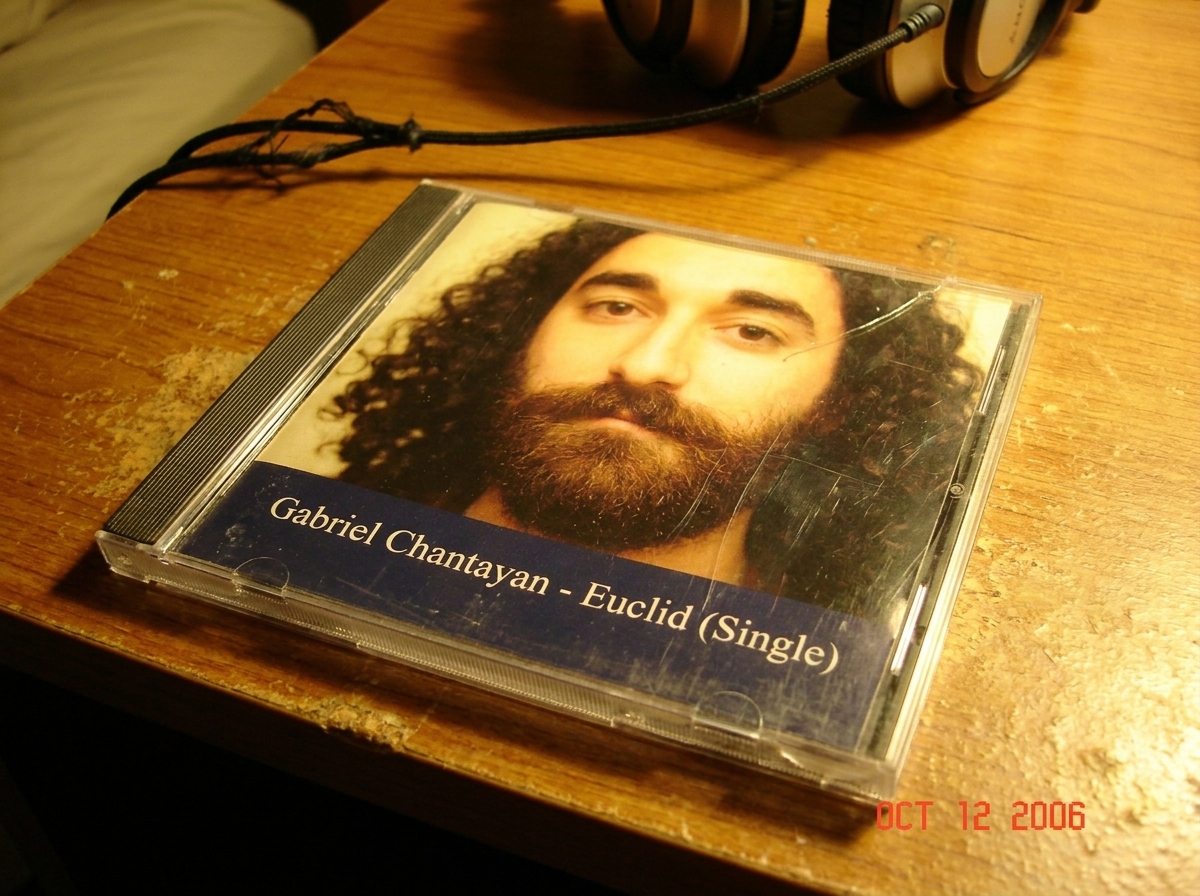 A close-up photo taken on a worn wooden desk of a used CD single in a jewel case. The label is scuffed and slightly torn, with 'Gabriel Chantayan - Euclid (Single)' printed in small white letters on a dark blue background. The case is cracked at the hinge. A pair of headphones with a frayed cord is 