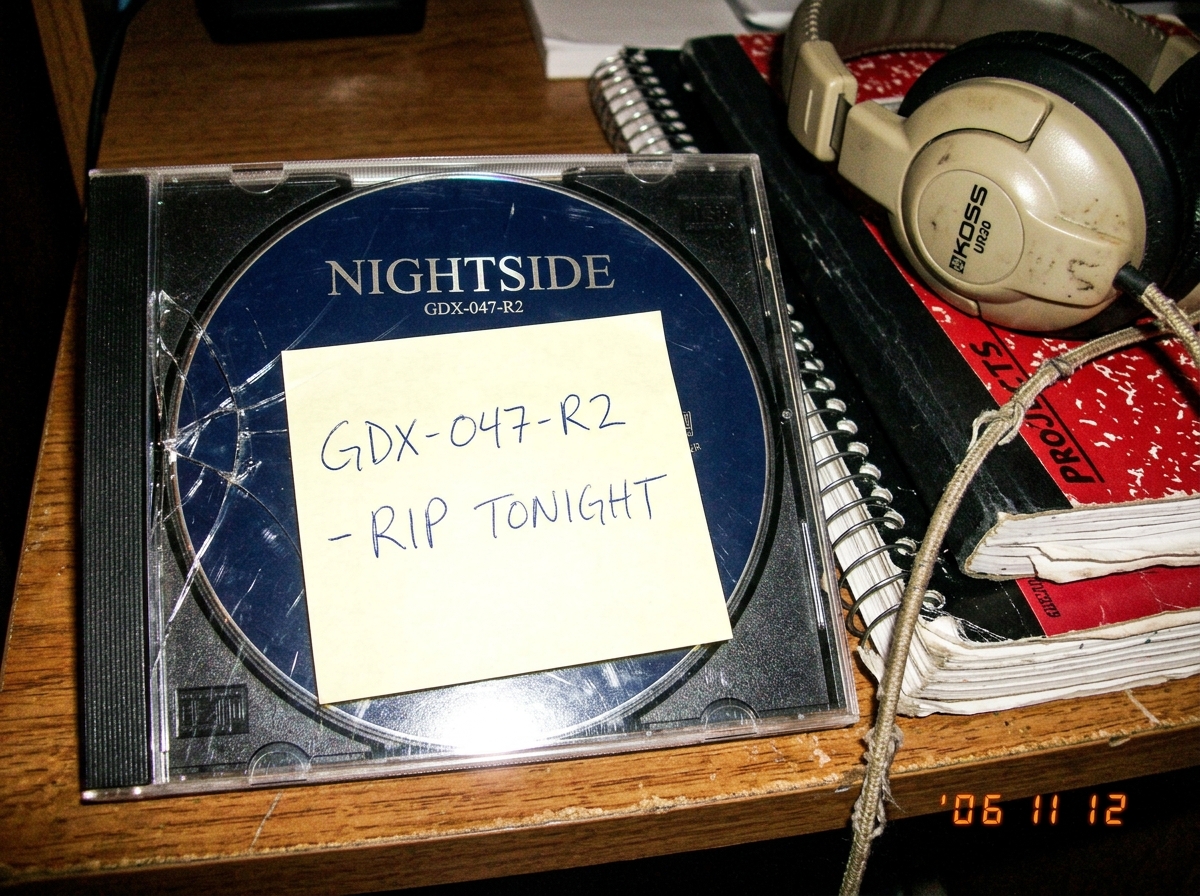 A close-up photo of a CD jewel case cracked at the hinge, with a handwritten sticky note on top reading 'GDX-047-R2 - RIP TONIGHT' in blue ballpoint pen. The CD itself is visible through the case, dark blue with silver text. A pair of beige headphones with a frayed cord sits partially in frame next