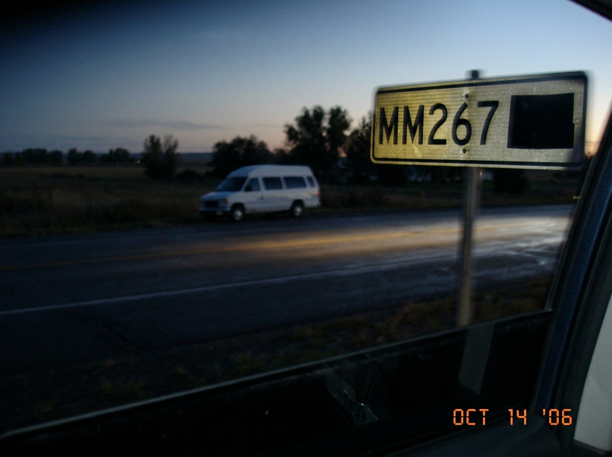 A blurry photo of a road sign that says 'MM267' with a Wyoming state outline next to it. In the background, a white church van is barely visible parked on the shoulder. The photo looks like it was taken from a moving car window at dusk.