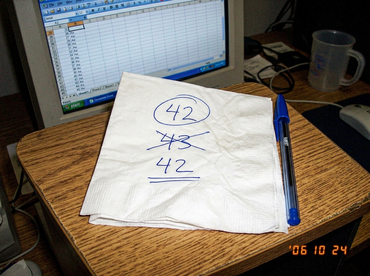 A folded napkin with handwritten numbers: '42' circled in blue ink, '43' crossed out, and '42' underlined. A pen sits next to it on a laminate desk surface. A computer monitor is partially visible in the background showing an Excel spreadsheet with columns labeled B2 through K47.