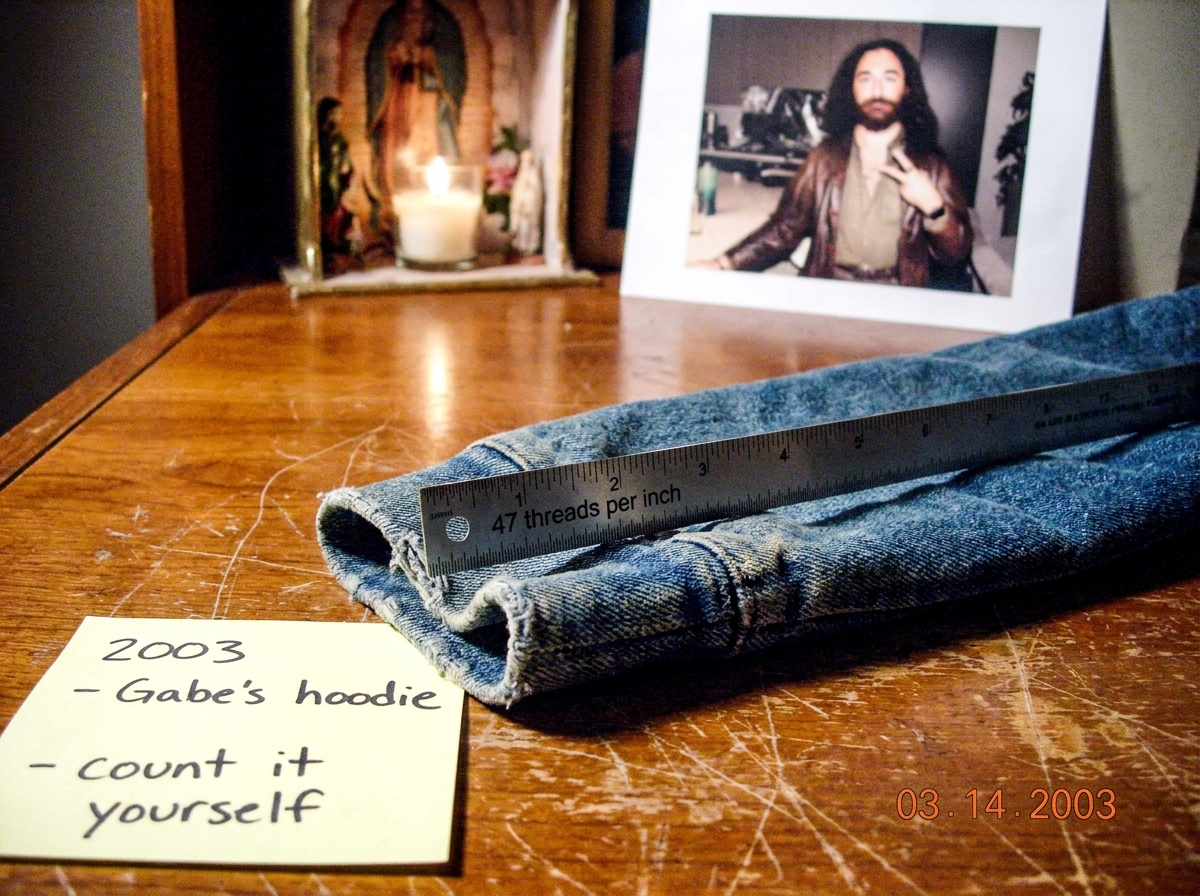 A close-up of a denim hoodie cuff with a ruler laid across it showing 47 threads per inch. The photo is slightly overexposed and taken on a worn wooden table. A handwritten sticky note at the edge reads '2003 - Gabe's hoodie - count it yourself'. In the background, a small shrine area with a candle 