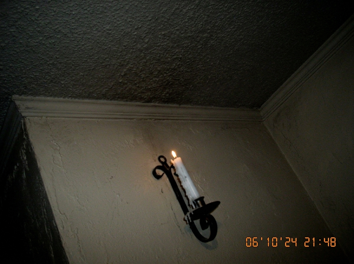 A slightly crooked wall sconce/candle holder on a beige wall, with a half-melted white candle in it. The flame is tiny and pathetic. The angle is badly composed, clearly taken on a 2006 flip phone camera - grainy, bad lighting, shows more of the ceiling than the candle.