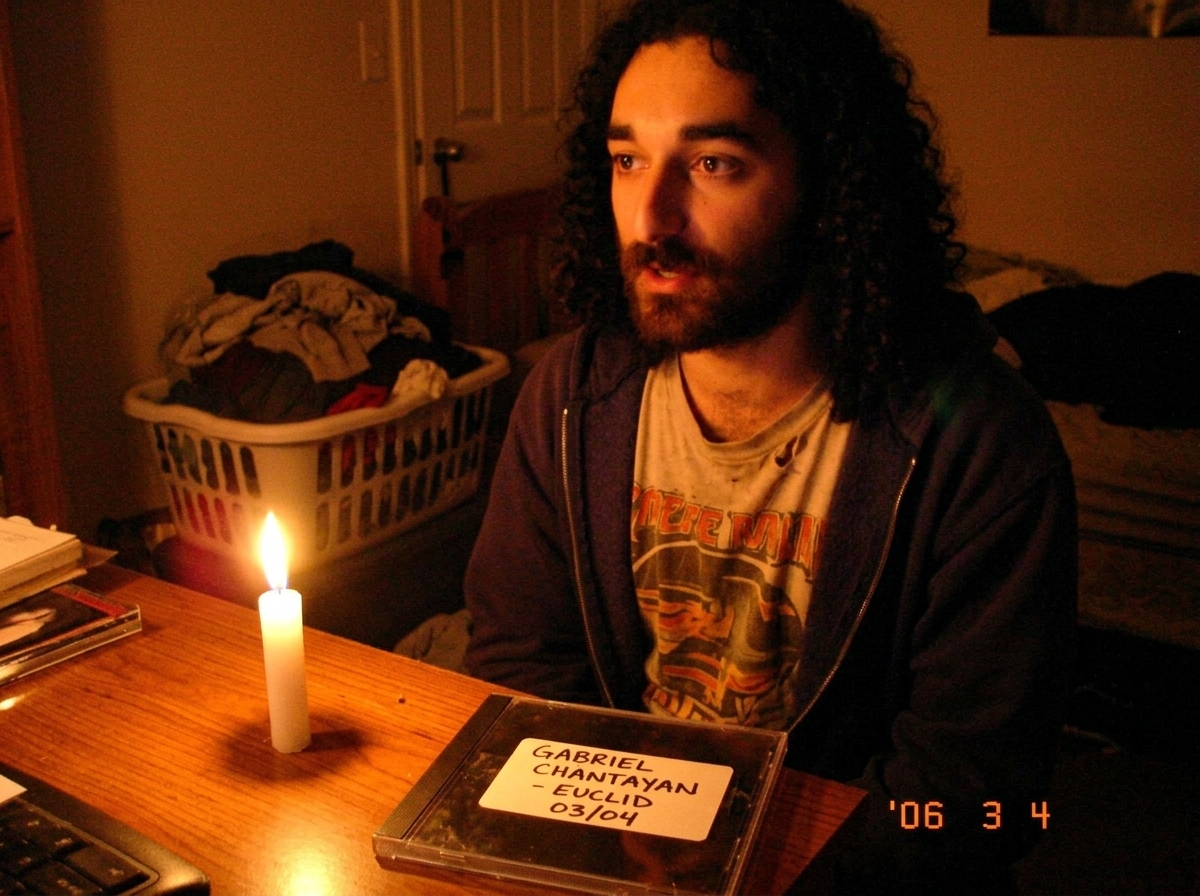 A grainy, dimly lit photo taken on a 2006 flip phone showing a single white candle on a cluttered desk next to a CD jewel case. The CD case has a worn, hand-labeled 'GABRIEL CHANTAYAN - EUCLID 03/04' sticker on it. The candle flame is small. In the background blur, a laundry basket full of folded cl