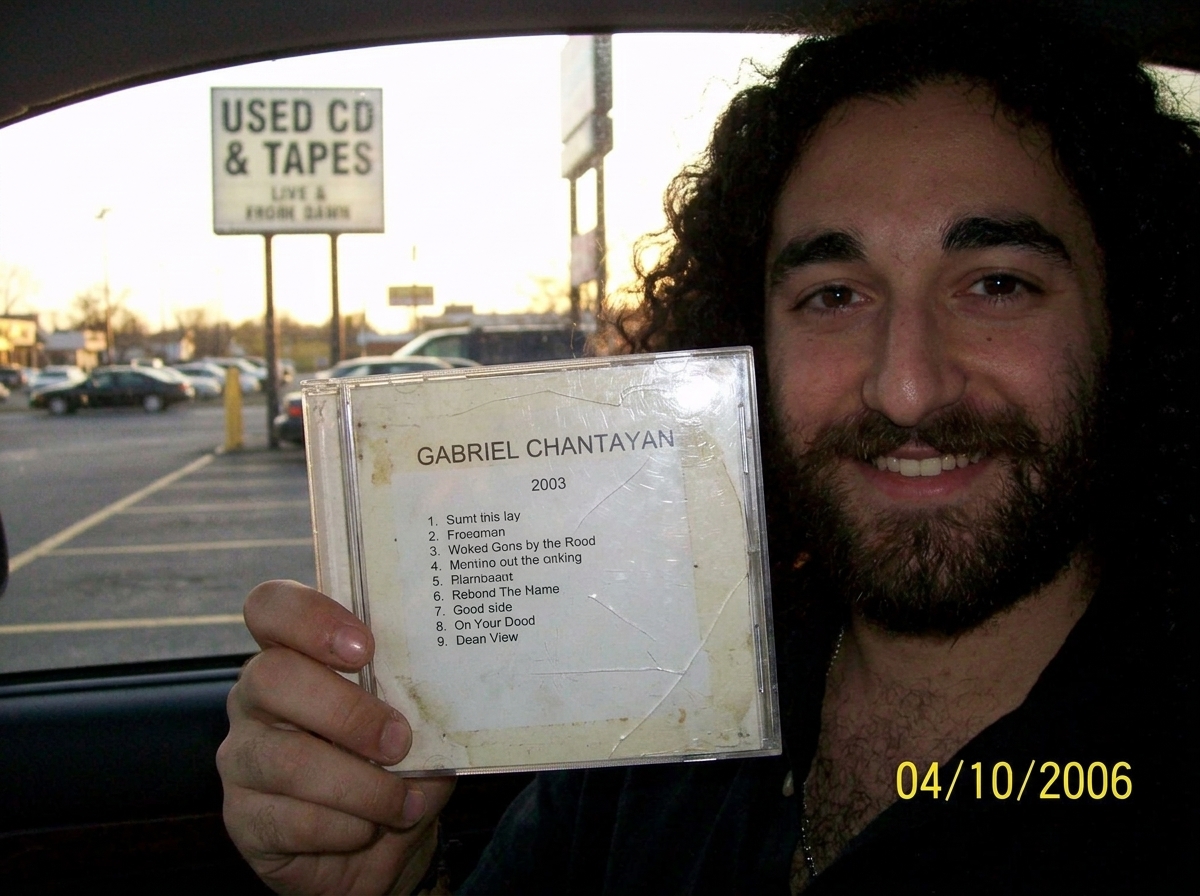 A worn CD single in a clear jewel case, held up against a late afternoon window. The label is heavily faded with age, the text barely readable - 'Gabriel Chantayan' in a sans-serif font with a track listing that appears to show a 2003 date. The edge of the disc has visible scuff marks and the case i