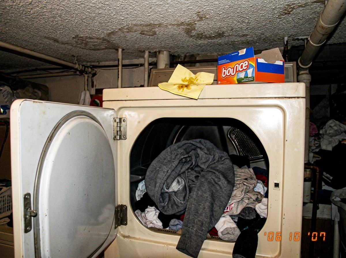 A grainy flip-phone photo of a clothes dryer door hanging open, with a single damp-looking hoodie visible inside on top of a pile of dry clothes. A crumpled taco truck napkin sits on top of the dryer next to a box of Bounce dryer sheets. The photo is poorly lit and clearly taken in a hurry, showing 