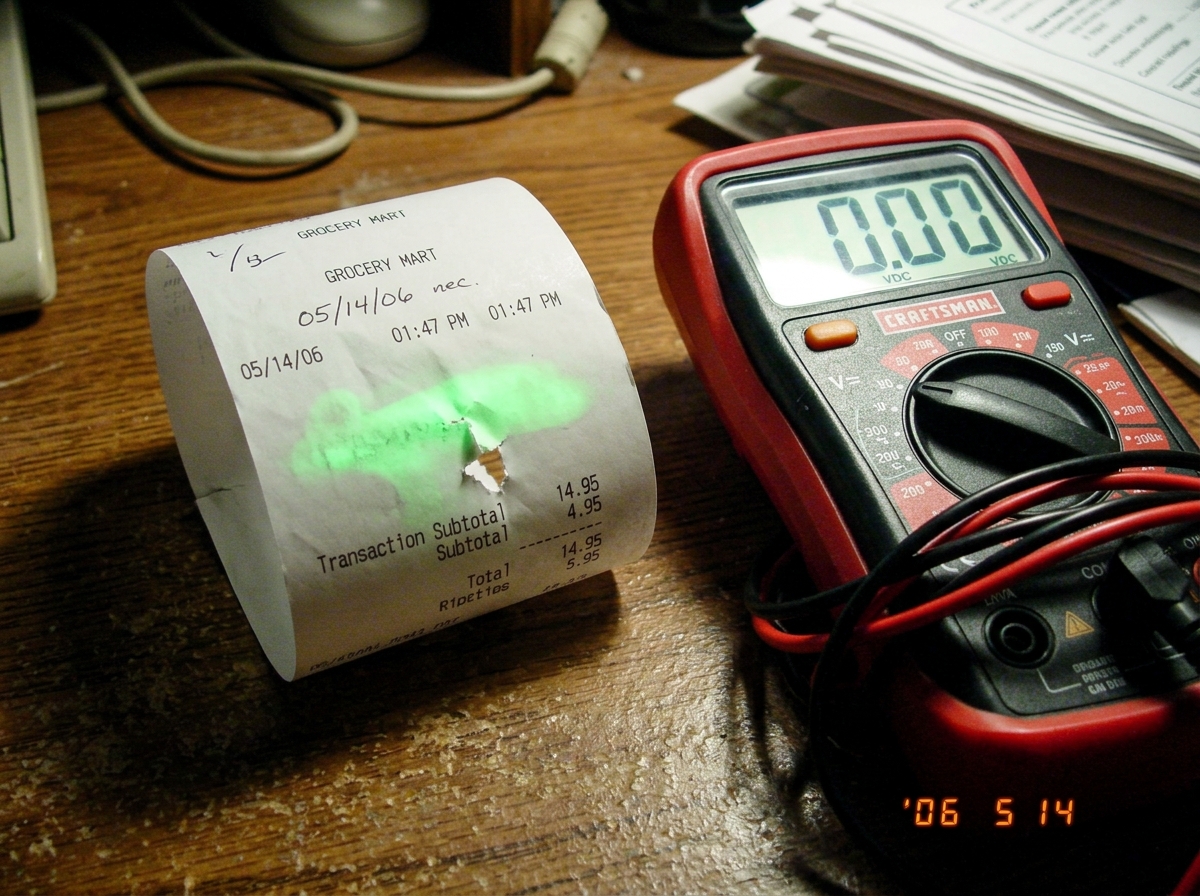 Thermal paper receipt showing green glow smudge at 1:47 timestamp with multimeter reading 0.00 resting beside it