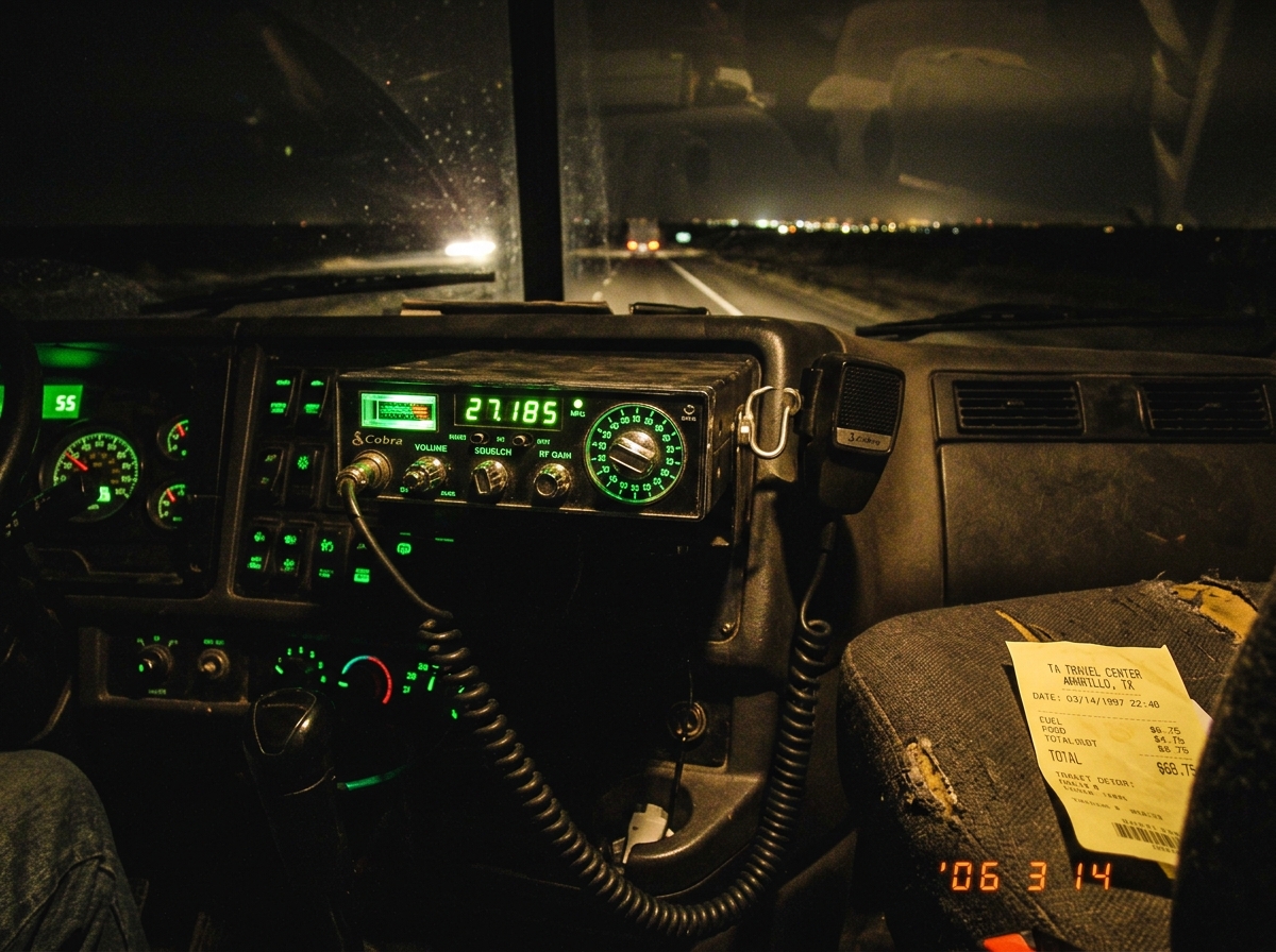 A CB radio mounted in a truck cab, the dashboard lit green, with the frequency dial visible around 27.185 and the mic hanging on its hook. A crumpled receipt with 'AMARILLO 1997' visible on the passenger seat. Night driving through desert visible through windshield.