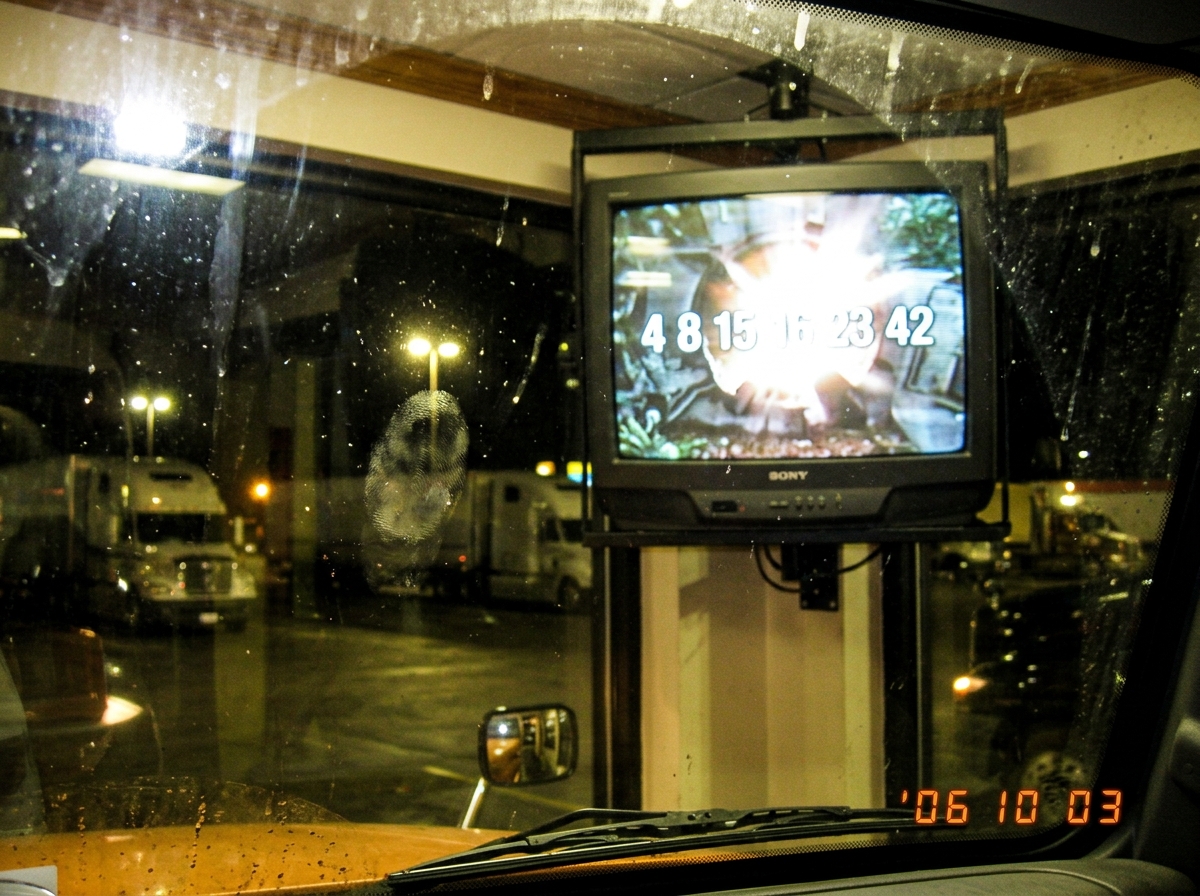 A blurry photo taken through a dirty truck windshield at night, showing a TV in a truck stop lounge with a frozen frame from Lost season 2 finale - the hatch door blown open, bright light pouring out, numbers 4 8 15 16 23 42 visible on the screen. The image has scanlines and a greasy thumbprint on t
