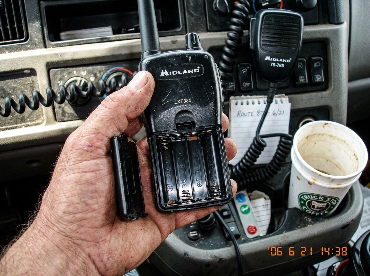A clear, well-lit photo of a two-way walkie-talkie held in a weathered hand, turned upside-down to show the battery compartment. The battery door is visibly open, exposing the empty battery slot and the metal contacts inside. The background is a truck dashboard with an empty coffee cup and a CB radi