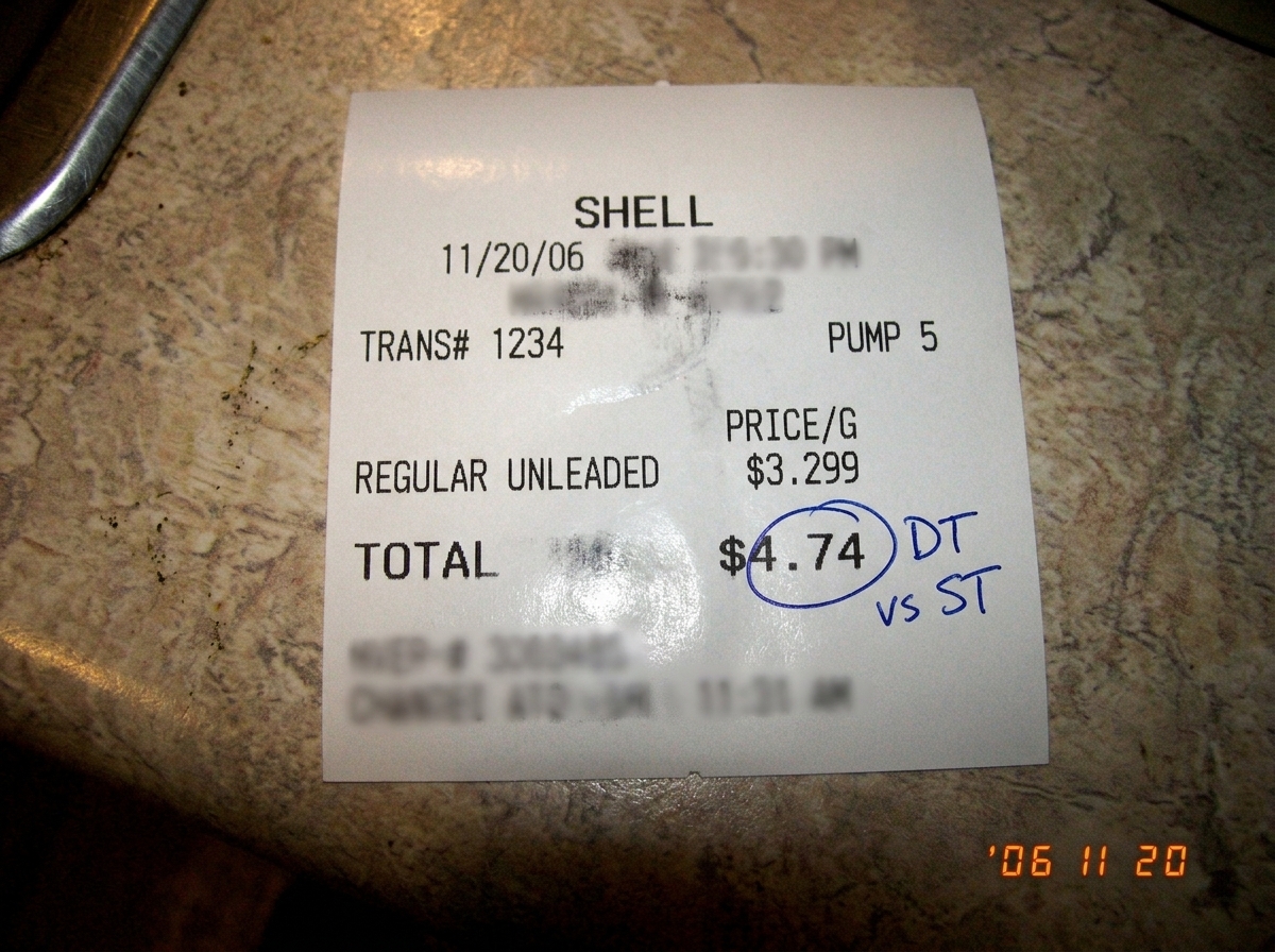 a blurry photo of a gas station receipt on a kitchen counter, the date is smudged and the total appears to be $4.74 - the 74 cent portion is circled in blue pen with a handwritten note "DT vs ST"