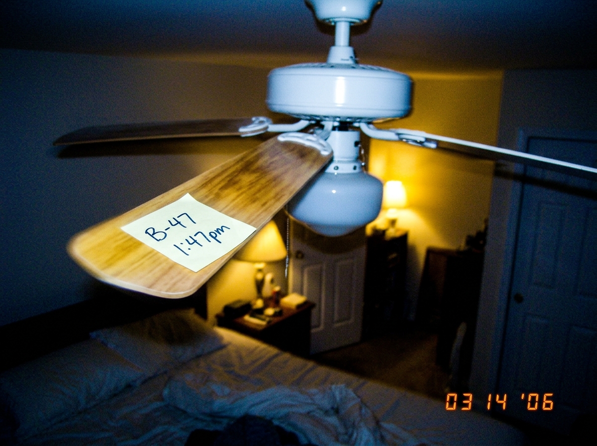 A blurry photo taken at night of a white ceiling fan with a wooden blade visible, a small handwritten sticky note stuck to the blade reads 'B-47 1:47pm', the room is dimly lit with yellow incandescent light