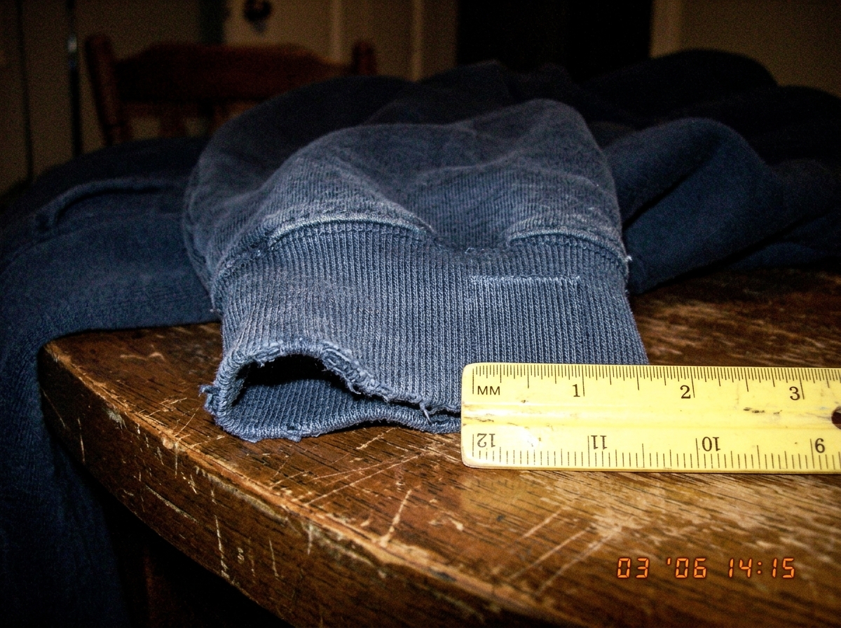 A close-up photograph of a dark blue hoodie cuff resting on a wooden table, showing the weave texture clearly. A small yellow ruler beside it measures the threads - 47 threads counted in one inch. The fabric is slightly faded, and the room lighting is warm and dim.