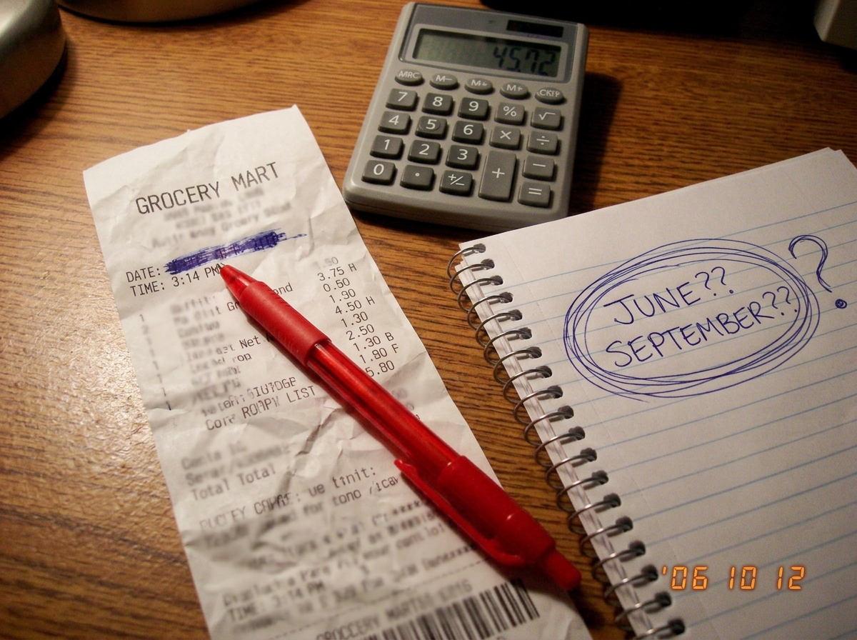A crumpled grocery receipt lying next to a calculator on a wooden desk. The receipt is partially unrolled, showing a timestamp of 3:14 but the date field is blurred/smeared. A red pen has been placed pointing at the date field. Next to the receipt is a spiral notebook with a hand-drawn question mark