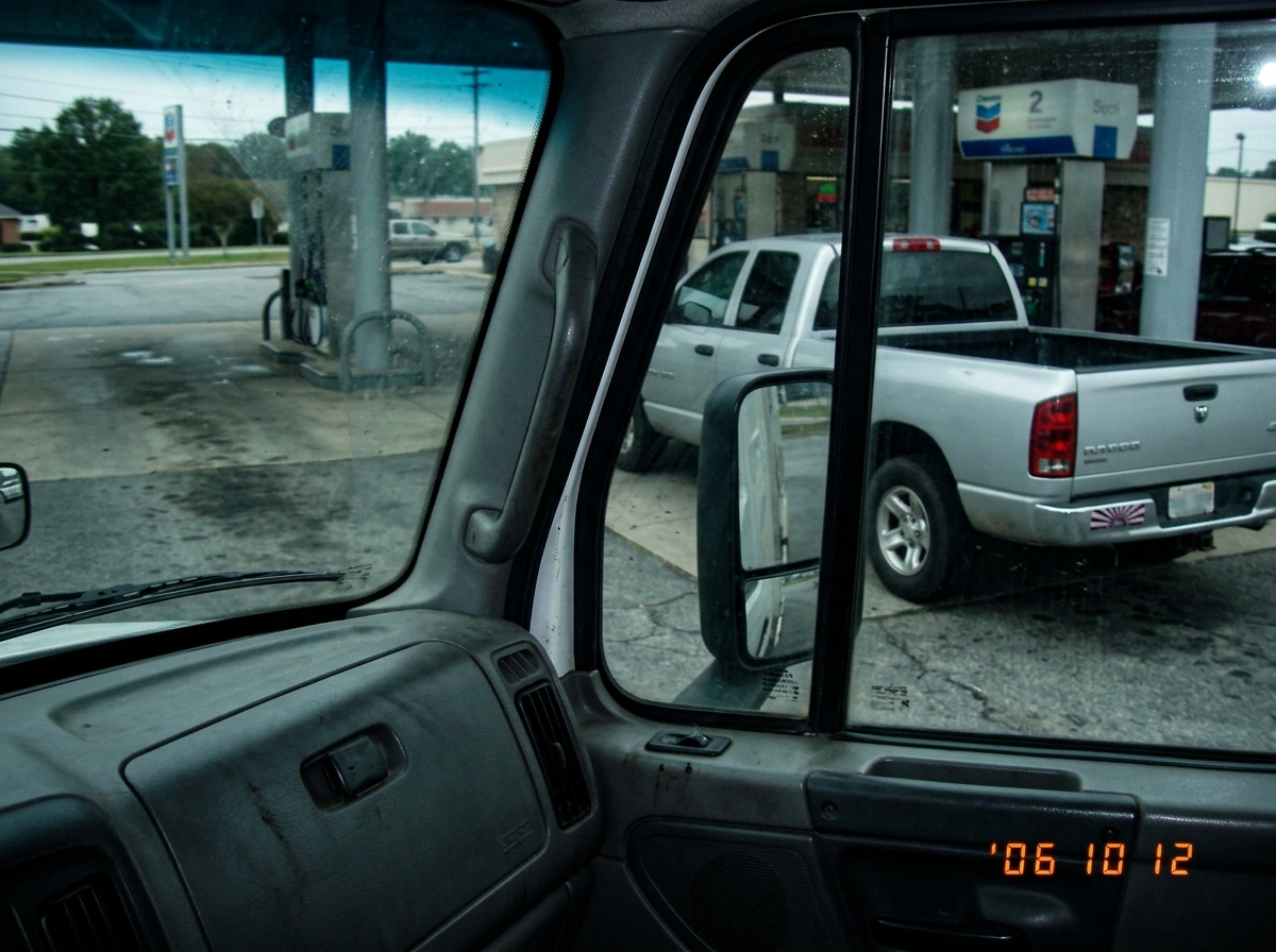 A blurry cell phone photo taken from inside a truck cab, showing a pickup truck in the next lane at a gas station. The truck has a bumper sticker that is difficult to make out, just a suggestion of radiating lines. The photo is poorly framed, mostly showing the truck's side mirror and a bit of the d