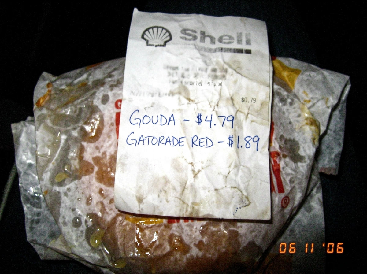 A crumpled gas station receipt with 'GOUDA - $4.79' and 'GATORADE RED - $1.89' handwritten on it in blue ballpoint pen, sitting on a greasy fast food wrapper, low-res camera phone photo taken at night with bad flash