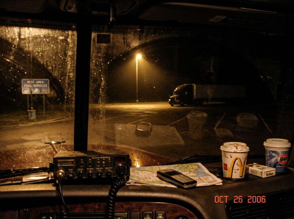 A blurry night photo taken from a truck cab, shot through the windshield at a rest area parking lot. The dashboard is visible at the bottom edge, cluttered with coffee cups and a CB radio. In the distance, a single lamppost casts a cone of yellow light onto an empty parking space. The photo has a gr
