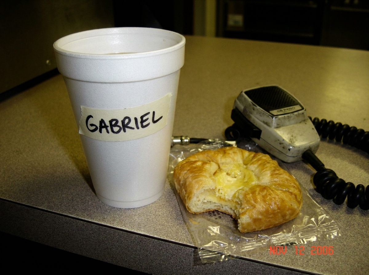 A blurry camera phone photo of a styrofoam coffee cup on a truck stop counter, with a hand-labeled piece of masking tape that says 'GABRIEL' in black sharpie on the side of the cup. In the background, a half-eaten gas station danish and a well-worn CB microphone.