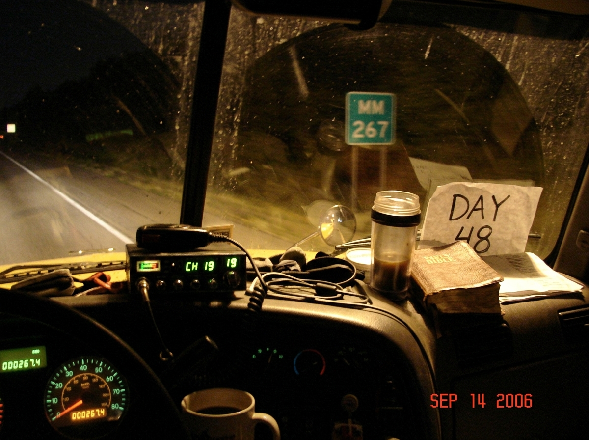 A blurry night photo taken from inside a semi truck cab, looking out the windshield. The dashboard shows a CB radio with the channel display lit up, and the odometer reads 267.4 miles. Through the windshield, a green highway sign is partially visible showing MM 267. In the passenger seat, a well-wor