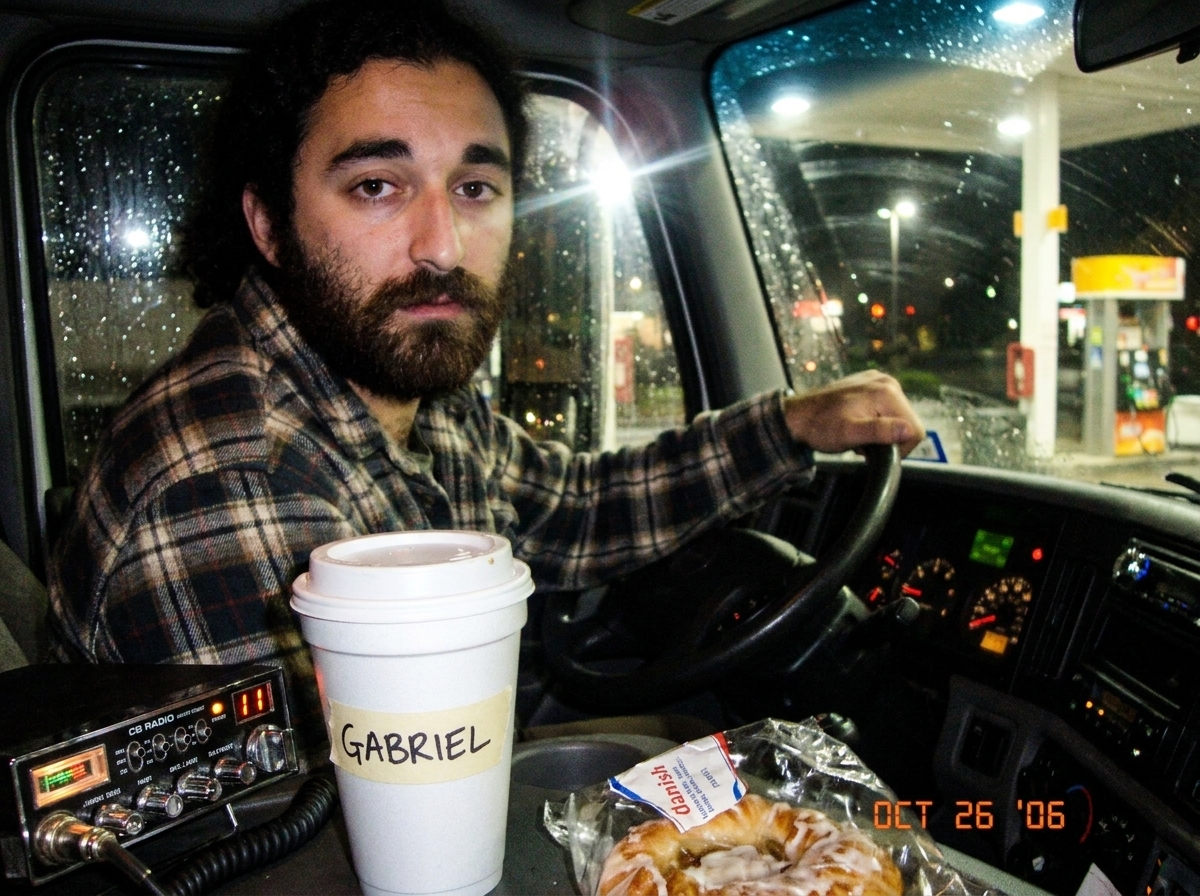 A blurry photo of a gas station coffee cup with masking tape labeled 'GABRIEL' on it, sitting next to a CB radio that shows channel 11. A danish wrapper is crumpled in the background. The photo is taken at night through a truck windshield with rain on the glass.