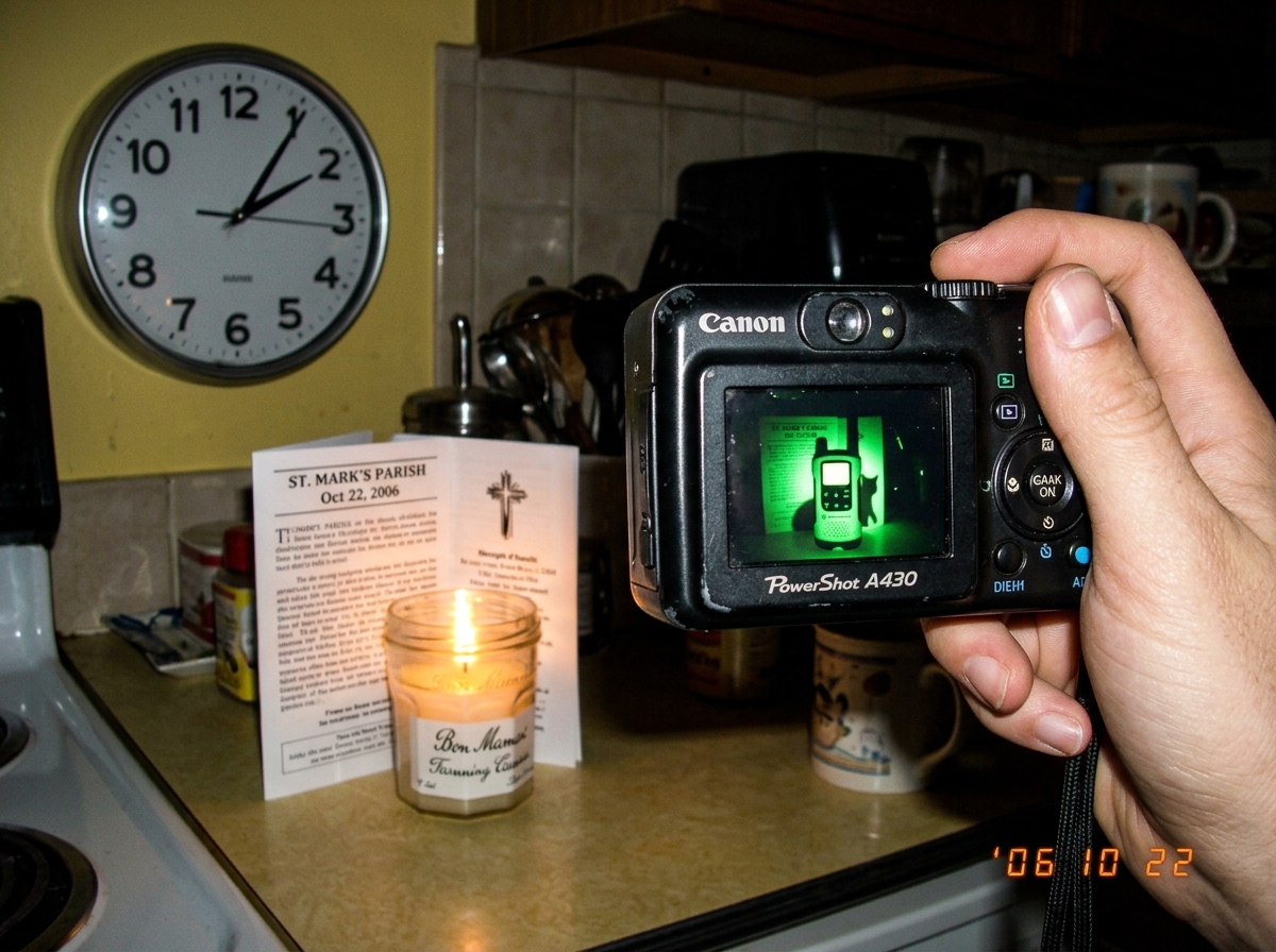 A hand holding an old black digital camera turned toward a small lit shrine on a kitchen counter: a jam jar with a candle, a folded church bulletin, and a round analog wall clock showing 1:47 in the corner. On the camera's rear LCD screen, a thumbnail of a green-glowing walkie talkie with a cat shad