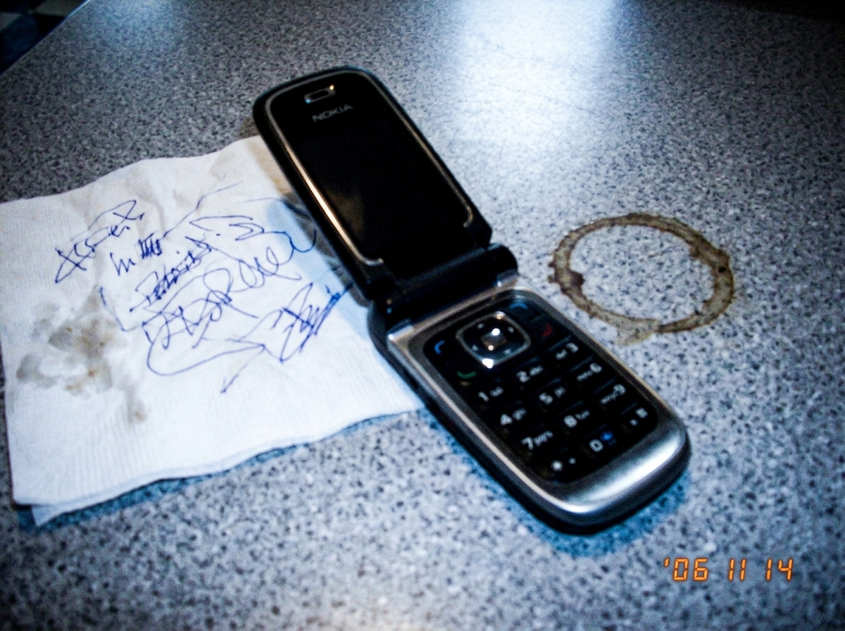 A blurry cell phone photo of a 2006-era Nokia flip phone next to a crumpled diner napkin with illegible blue ink scribbles, on a Formica tabletop with a coffee ring visible. Taken at night with flash.