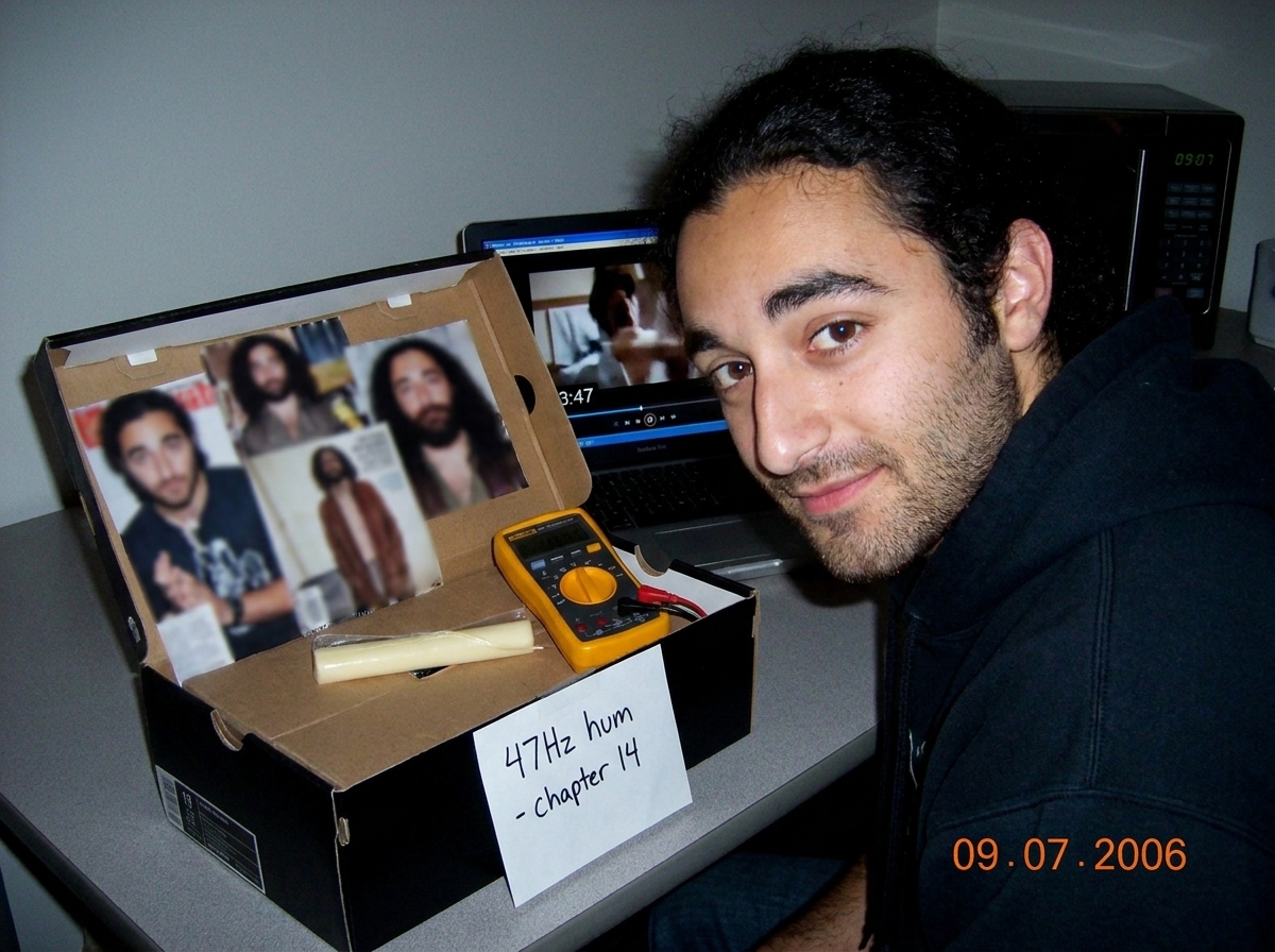 My shoebox altar with Gabriel magazine clippings, blurry teen magazine photo, string cheese, tiny Fluke multimeter, handwritten note '47Hz hum - chapter 14', a laptop showing a paused frame at 3:47, and a microwave clock reading 09-07