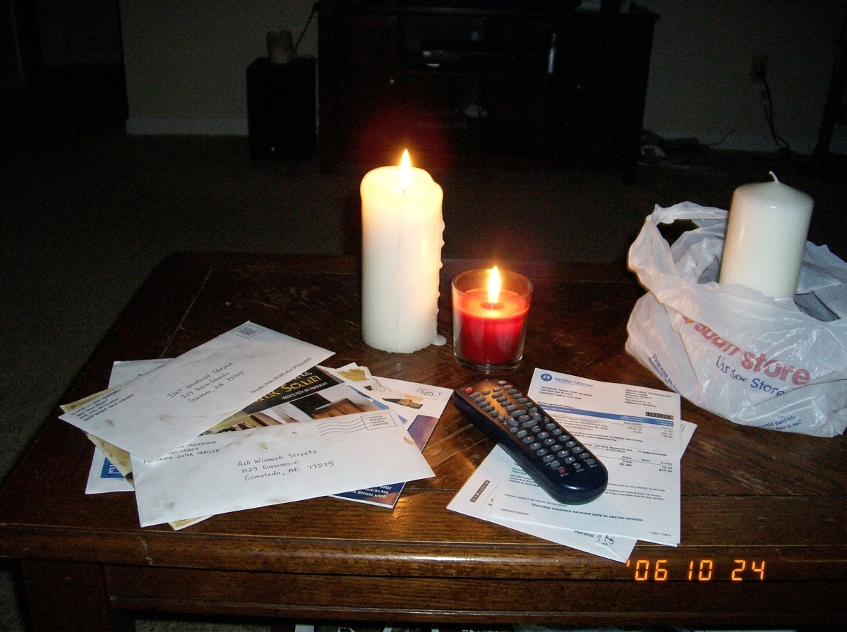 A blurry, low-light photo taken with a flip phone of two lit candles on a wooden coffee table - one tall white pillar, one short red votive in a glass holder - surrounded by discarded mail and a TV remote. The flames are slightly out of focus and the image grain is heavy. A third unlit candle sits o