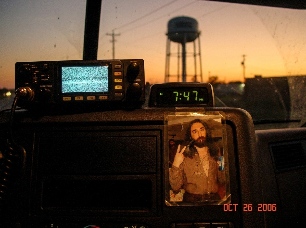 A photo of a CB radio's frequency display set to a static-filled channel, taken from inside a truck cab at dusk. The dashboard shows a digital clock reading 7:47 PM. In the background through the windshield, a water tower is silhouetted against an orange sunset sky. A small laminated photo of Gabrie