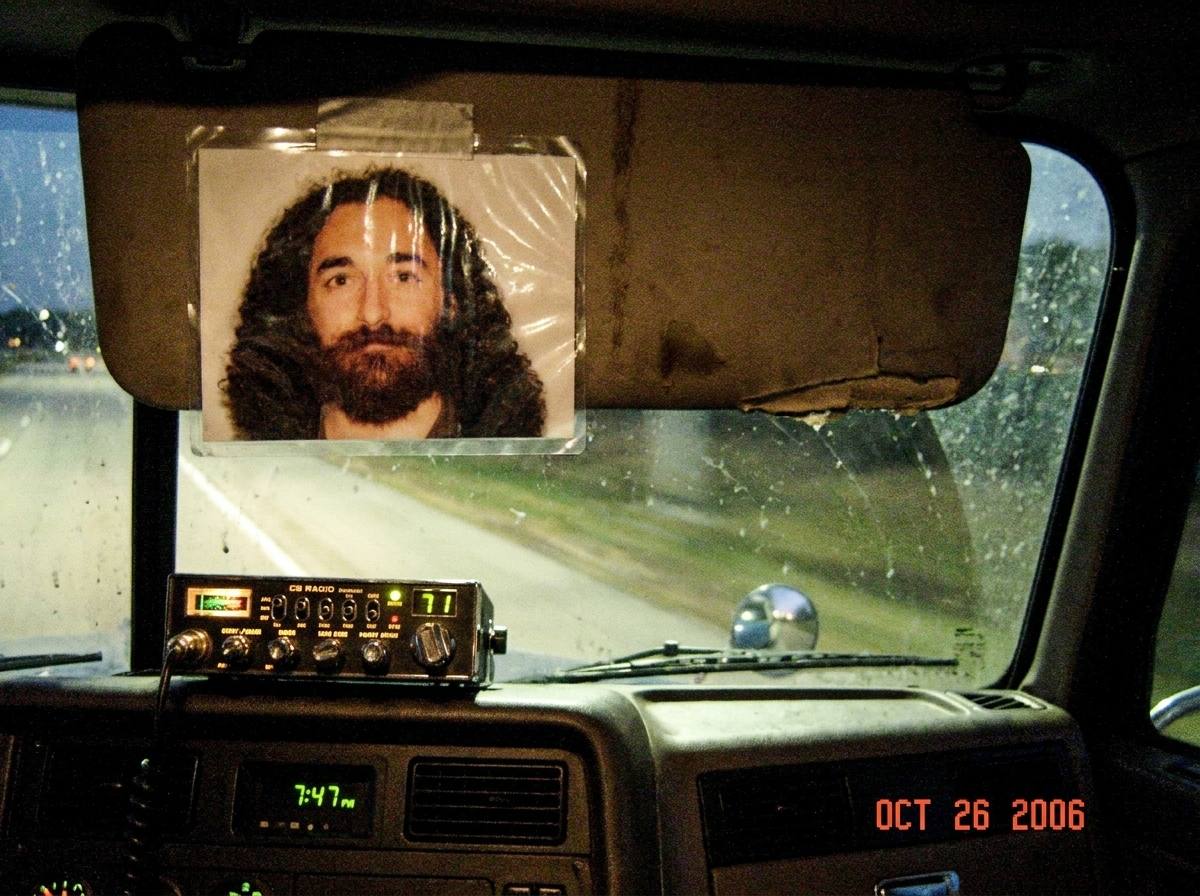 A laminated photo of Gabriel Chantayan taped to a semi truck's sun visor, with a CB radio and a dashboard reading 7:47 PM visible in the background through the windshield