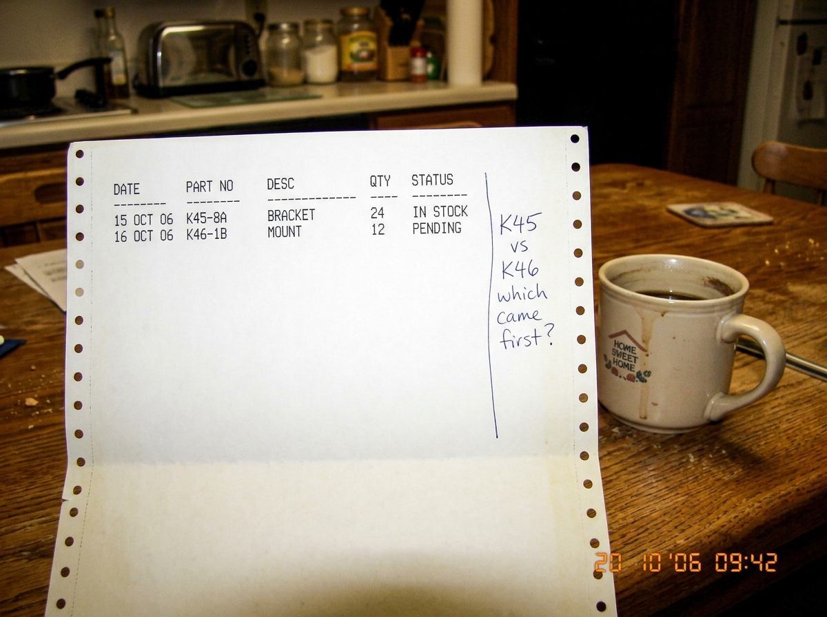 A photo of a dot matrix printer output sheet on a kitchen table, with a handwritten note in blue ink in the margin reading 'K45 vs K46 which came first?' next to a coffee cup