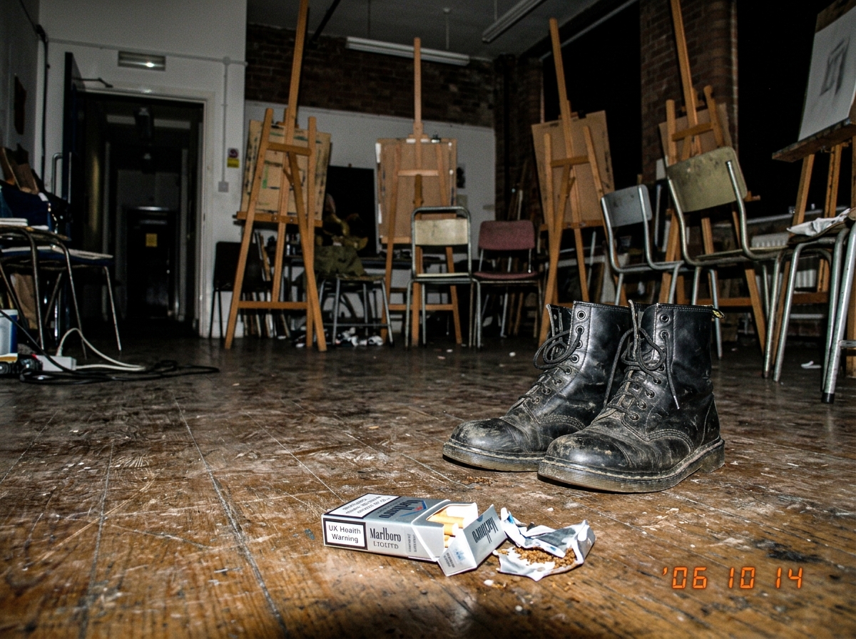 A blurry phone photo of an art studio with easels and critique chairs arranged in a semicircle. The focus is on the floor, showing a pair of worn black boots standing at the edge of the circle, slightly turned toward the door. A half-unrolled cigarette packet is visible on the floor nearby.
