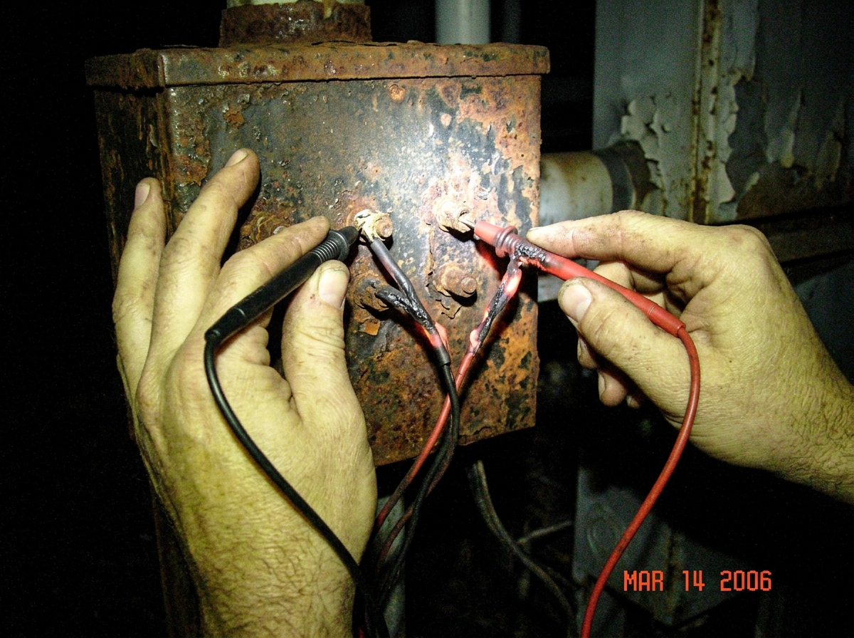 hands pressed against a rusted metal utility box with multimeter probes clamped between fingers and terminal posts, the probe insulation appears melted and glowing faintly orange