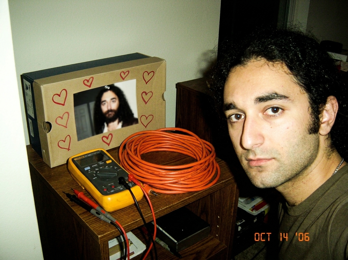 small shrine desk setup with a tiny blurry photo of Gabriel in a shoebox, drawn hearts around edges, a Fluke multimeter and coiled 16awg extension cord beside it