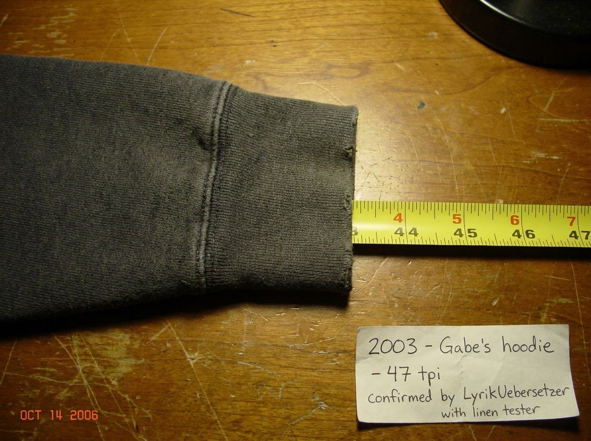 A close-up photo of a worn fabric cuff from a hoodie, laid flat on a wooden surface next to a measuring tape with inch markings. The weave is clearly visible, threads are countable at approximately 47 per inch. A handwritten paper label in the corner of the photo says '2003 - Gabe's hoodie - 47 tpi