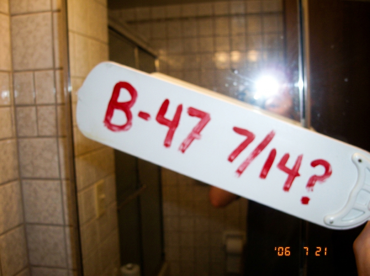 A ceiling fan blade photographed in a bathroom mirror selfie style, spray-painted with 'B-47 7/14?' in red. The blade is white, the bathroom is tiled, and the photo is blurry.