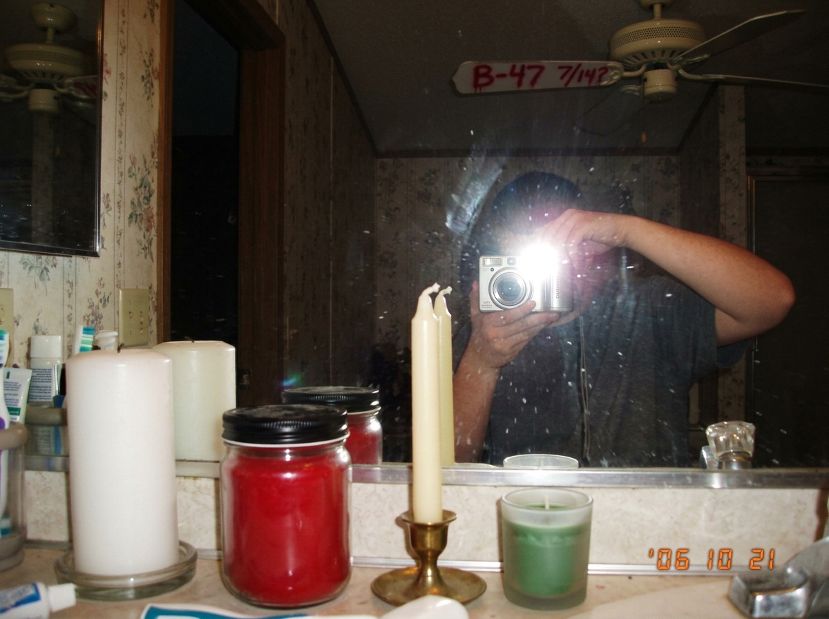 A blurry bathroom mirror selfie showing a bathroom counter with four unlit candles - white pillar, red jar candle, cream taper, and a short green votive - arranged in a row. In the mirror reflection, you can see someone's arm holding a phone and a partially visible ceiling fan blade behind them spra