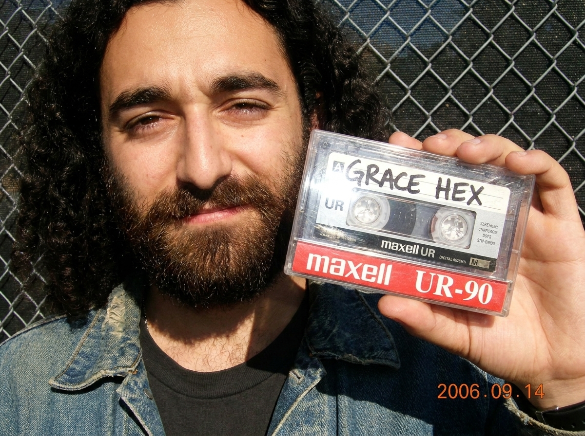 A worn, scuffed Maxell UR-90 cassette tape held in two hands in bright sunlight. The handwritten label says 'GRACE HEX' in black permanent marker. The tape's clear shell is smudged and the visible oxide reel is slightly uneven. The photo is taken outdoors at a slight angle, with chain-link fence vis