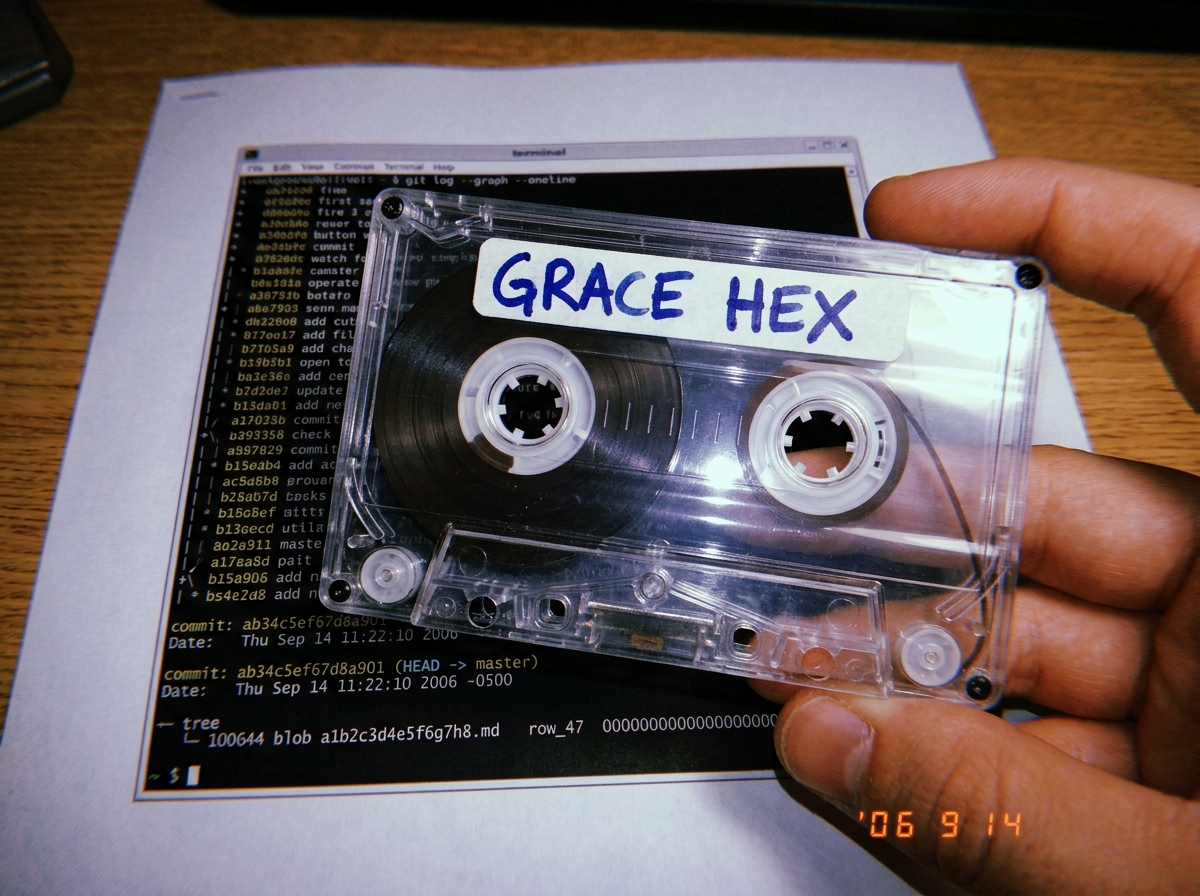 A close-up photo of a cassette tape's transparent shell held over a printed git log page. The tape's oxide reel is visible through the plastic, and the handwritten 'GRACE HEX' label is crisp in the frame. Underneath the tape, a terminal window screenshot shows 'ls-tree' output with a line reading 'r