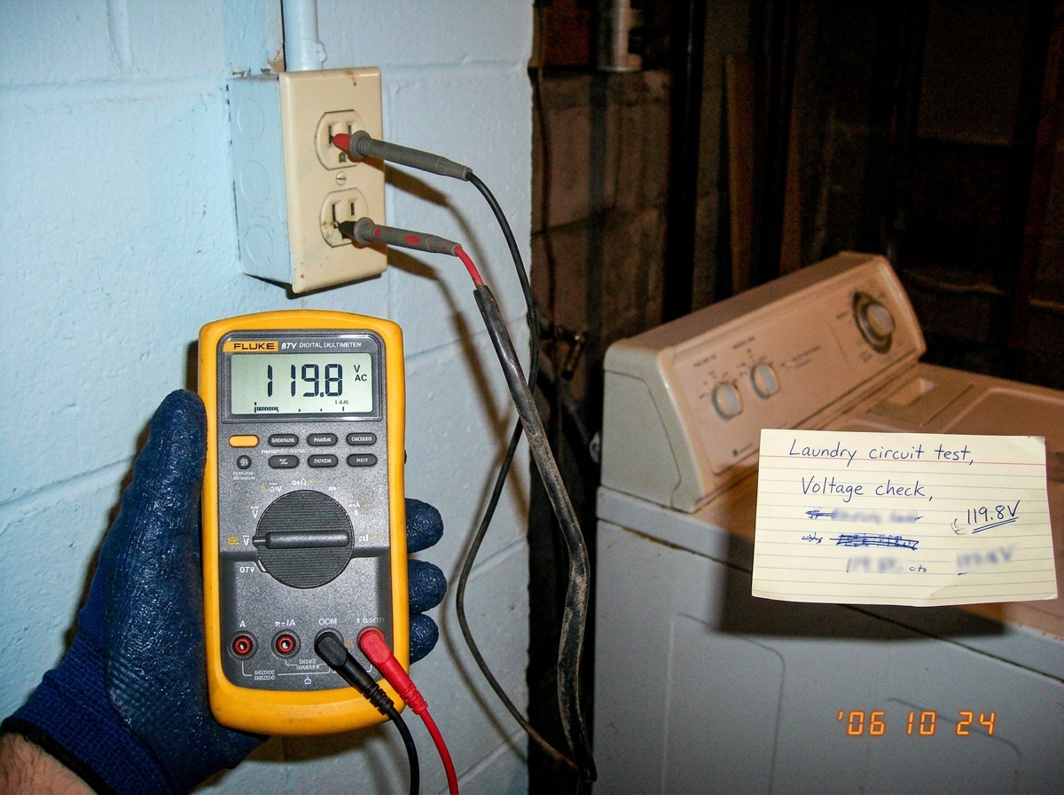 A Fluke 87V multimeter held in a gloved hand with probes in a wall socket in a basement laundry room. Reading 119.8V. Index card with handwritten notes on a dryer in the background.