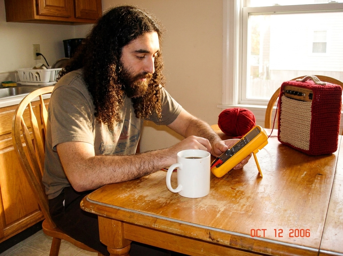 A yellow Fluke 87V digital multimeter sitting on a wooden kitchen table next to a white ceramic coffee mug, with a ball of red yarn and a nearly-finished crocheted Gabe-amp in the background under warm afternoon light streaming through a window