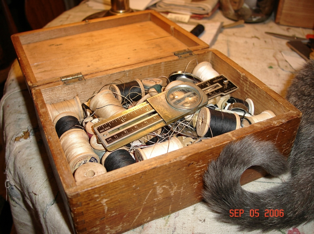 A close-up photo taken indoors under warm incandescent light showing a worn wooden sewing box with its lid open. Inside, resting on a tangle of old spools of thread (most are beige, black, and white), lies a vintage sewing gauge with a brass slider and a small circular magnifying lens built into the